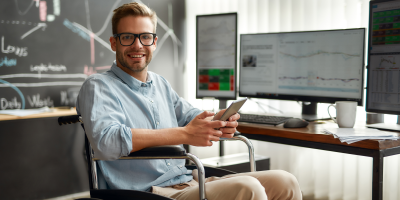 Portrait of young and cheerful male businessman in a wheelchair holding his smartphone and smiling while sitting at his workplace in modern office. Handicapped worker. Trading online. Stock market - leadership strategies for wellness